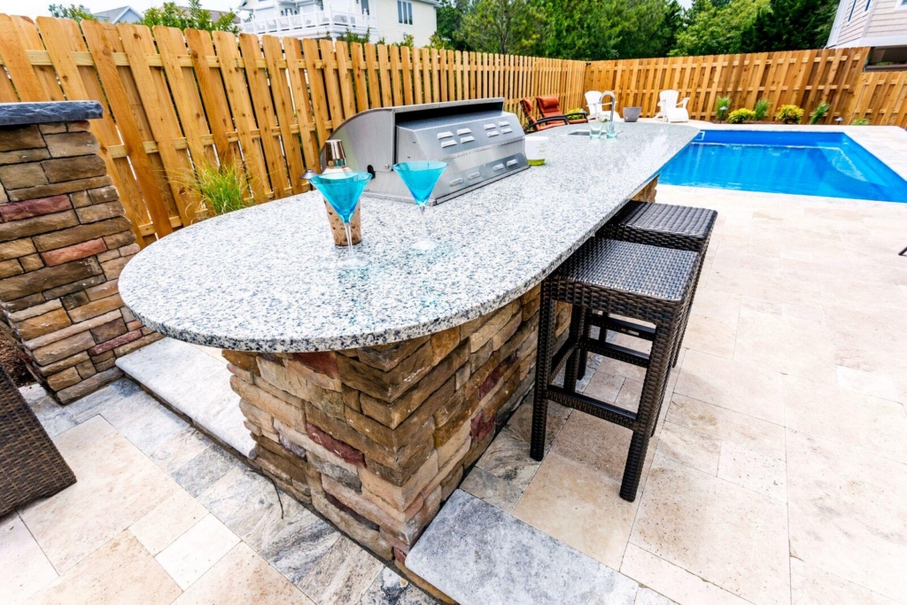Outdoor granite bar with two blue cocktails, wicker bar stools, and a built-in grill beside a luxury pool, surrounded by a wooden fence—ideal for enjoying outdoor kitchens in style.
