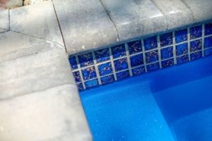Close-up of a swimming pool edge showing blue water, square blue tiles, and concrete coping with visible wear—an inviting detail often found in luxury pools or custom spas. Some debris is visible on the tile grout.