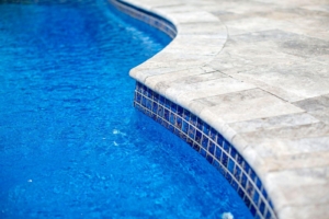 Close-up of the edge of a luxury pool with blue water and light-colored stone tiles, perfect for complementing custom spas or outdoor kitchens in your backyard oasis.