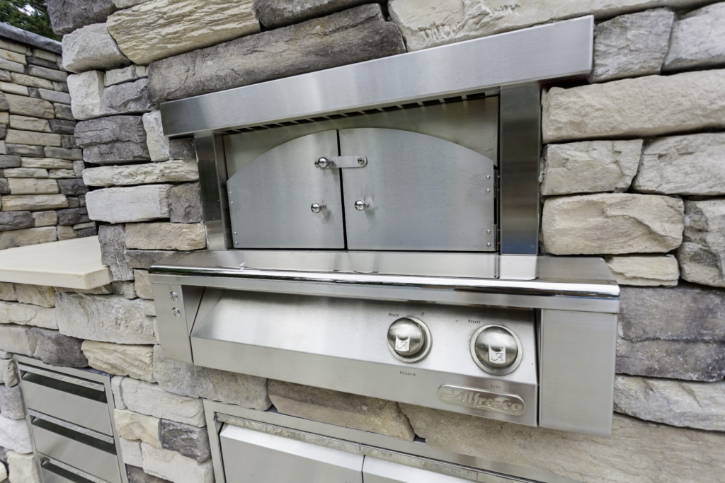 A stainless steel outdoor pizza oven with closed double doors and two control knobs, built into a stone wall—an elegant addition to outdoor kitchens.