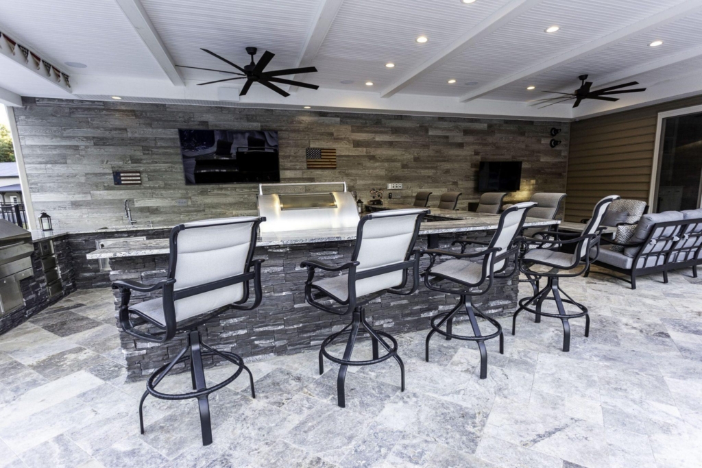 Modern outdoor kitchen with a stone bar, five barstools, built-in grill, TV, ceiling fans, and tiled floor—perfect for outdoor living and entertaining by your fiberglass pool.