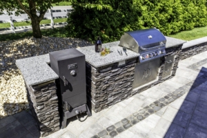 Outdoor stone kitchen with a built-in grill, smoker, granite countertops, and drinks on the counter—set on a patio with greenery, a white fence, and views of luxury pools in the background.