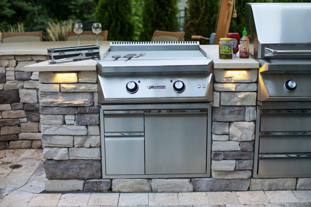 Outdoor grill station with stainless steel grill, utensils on top, and condiments nearby, built into a stone counter with cabinets below—perfect for enhancing your outdoor living space.