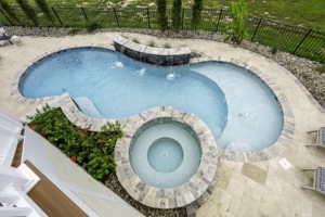 Aerial view of a backyard pool with a connected round hot tub, waterfall feature, surrounding stone patio, and plenty of space for outdoor living—perfect for relaxing or entertaining by your stunning fiberglass pool.
