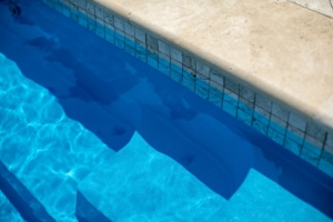 Close-up of blue swimming pool steps leading into clear water, next to a beige tiled pool edge—an inviting entryway often found in luxury pools designed for elevated outdoor living.