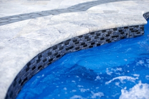 Close-up of the edge of an inground pool with blue water, textured stone tiles, and a decorative tile border.