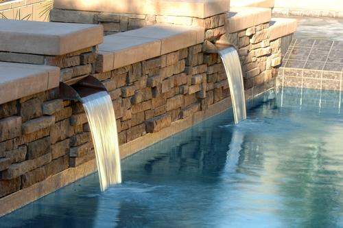 A stone wall with two built-in spouts pours water into an inground pool, creating small waterfalls that enhance the beauty of outdoor living.