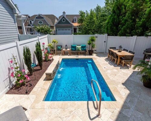 Small backyard with a rectangular luxury pool, lounge chairs, potted plants, flowers, and a patio dining set on stone tile flooring, enclosed by a white fence.