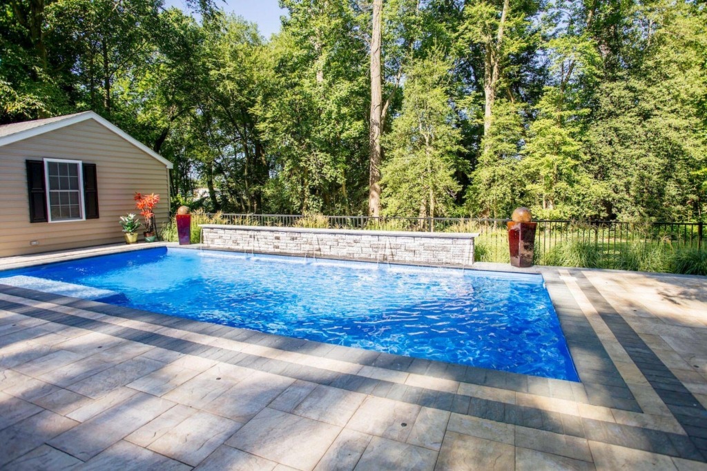 Rectangular luxury pool beside a small beige building, surrounded by trees and a stone-tiled patio—perfect for relaxing or entertaining by the pool.
