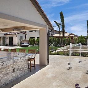Outdoor patio area with a stone bar and wooden stools overlooks a landscaped backyard featuring lush grass, trees, a modern fountain, and luxury pools—all set beneath a clear blue sky for the ultimate outdoor living experience.