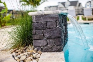 Close-up of a stone water feature pouring into a swimming pool, surrounded by decorative rocks and tall grass. Luxury pools, custom spas, and outdoor kitchens complement the stylish houses and patio furniture in the background.