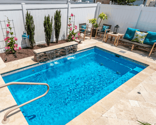 Rectangular backyard pool with a small stone waterfall, surrounded by tile decking, patio furniture, potted plants, and a white privacy fence—ideal for pairing fiberglass pools with custom spas for added relaxation.