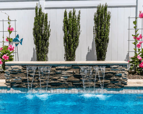 A backyard fiberglass pool with blue water, a stone waterfall feature, three tall shrubs, pink flowers, and a white privacy fence creates a perfect outdoor living retreat.