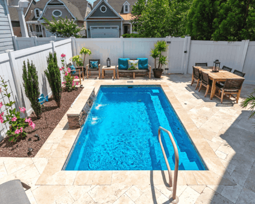 A small backyard features a rectangular inground pool, lounge chairs, a dining table, potted plants, and surrounding beige tile flooring—perfect for stylish outdoor living.