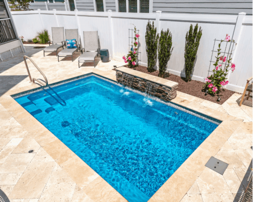 Rectangular inground pool with clear blue water, a waterfall feature, three lounge chairs, and flowering plants along a white fence—perfect for those seeking luxury pools in their backyard retreat.