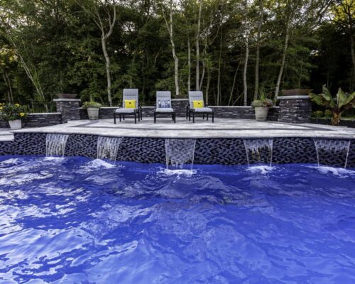 Three lounge chairs and two potted plants sit on a stone patio behind a fiberglass pool with cascading water features; this inviting outdoor living space is framed by trees in the background.