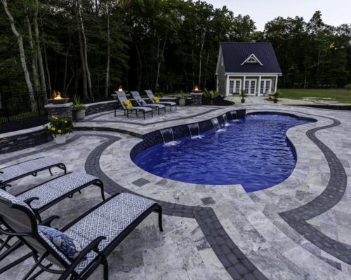A stone patio surrounds a curvy fiberglass pool with built-in fountains, lounge chairs, potted plants, and a small house in the background, bordered by trees—perfect for relaxing or entertaining with outdoor living at its finest.