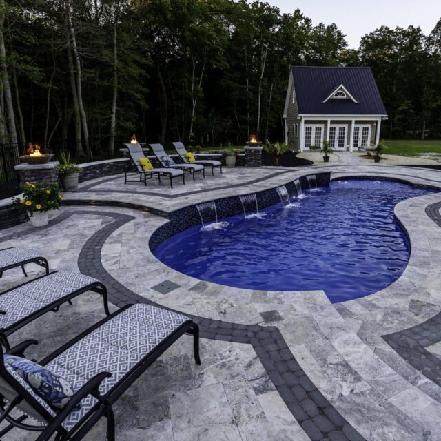 A stone patio surrounds a curvy fiberglass pool with built-in fountains, lounge chairs, potted plants, and a small house in the background, bordered by trees—perfect for relaxing or entertaining with outdoor living at its finest.