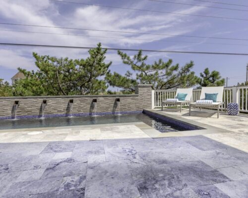 Outdoor swimming pool with five water spouts, a stone tile deck, lounge chairs with blue cushions, and trees in the background under a partly cloudy sky—perfect for those who love elegant inground pools.
