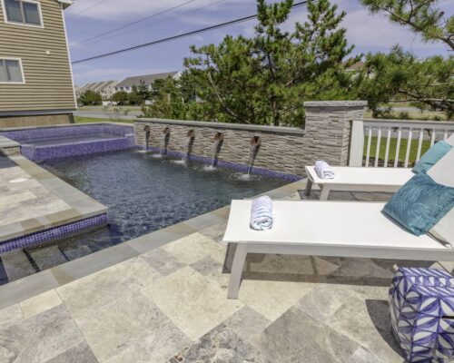 Modern backyard pool with water spouts, two white lounge chairs with blue accents, rolled towels, and a tiled stone patio beside a landscaped area—perfect for outdoor living or complementing fiberglass pools.
