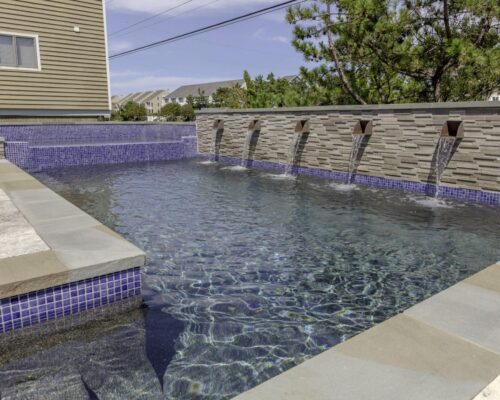 Rectangular backyard inground pool with blue tile accents, stone edging, and four water spouts built into a textured wall—perfect for refined outdoor living.