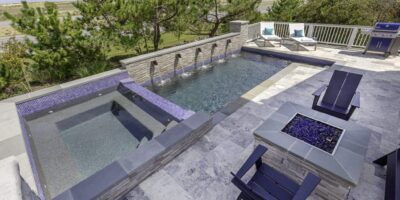 Modern backyard featuring an inground pool with attached custom spa, stone patio, fire pit, outdoor kitchen with grill, and two lounge chairs; trees and distant buildings in the background.