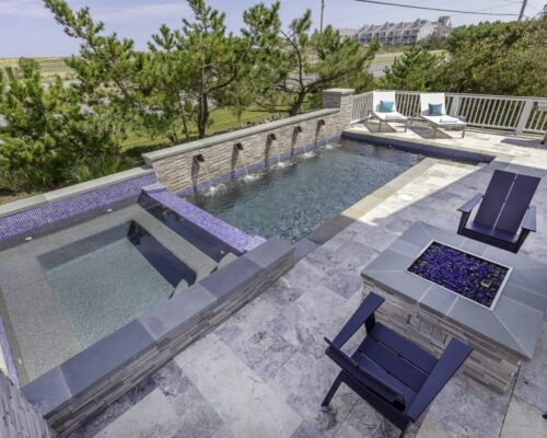 Modern backyard featuring an inground pool with attached custom spa, stone patio, fire pit, outdoor kitchen with grill, and two lounge chairs; trees and distant buildings in the background.