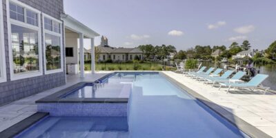 Modern backyard with an inground rectangular swimming pool, attached hot tub, and several blue lounge chairs on a white pool deck near a waterfront—perfect for relaxing and enjoying outdoor living.