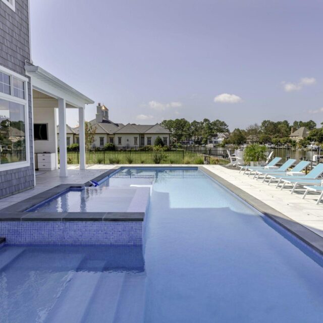 Modern backyard with an inground rectangular swimming pool, attached hot tub, and several blue lounge chairs on a white pool deck near a waterfront—perfect for relaxing and enjoying outdoor living.