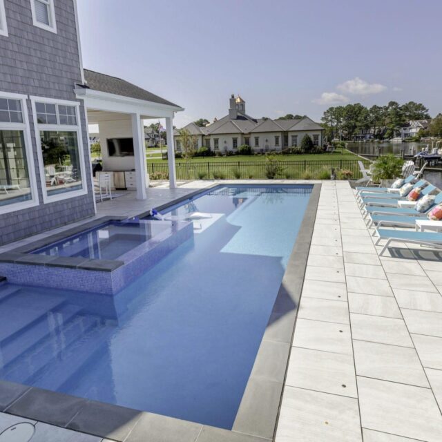 Modern backyard with a rectangular inground pool, custom spa hot tub, lounge chairs with colorful pillows, and waterfront view next to a gray house—perfect for outdoor living.