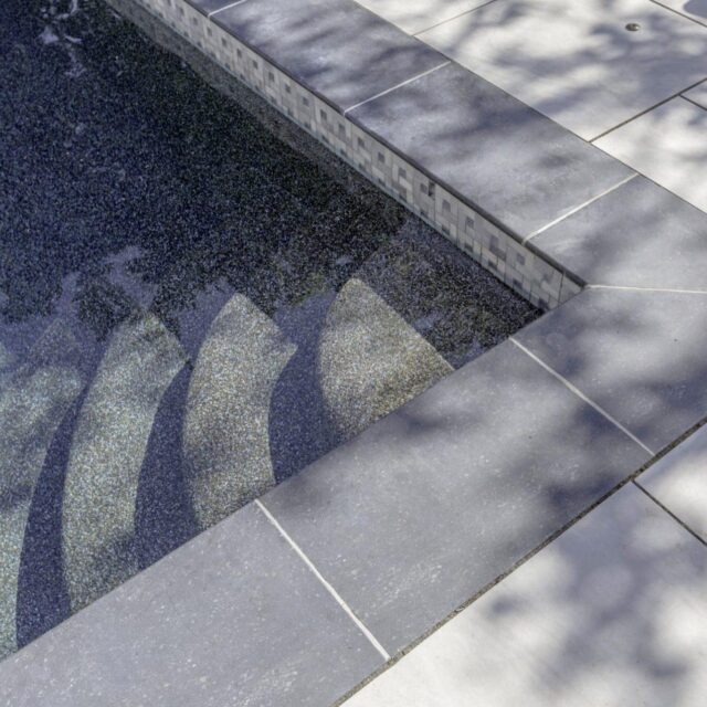 Shallow steps lead into a tiled inground pool with sunlight and tree shadows dancing on the surrounding light-colored pool deck, creating a perfect setting for outdoor living.