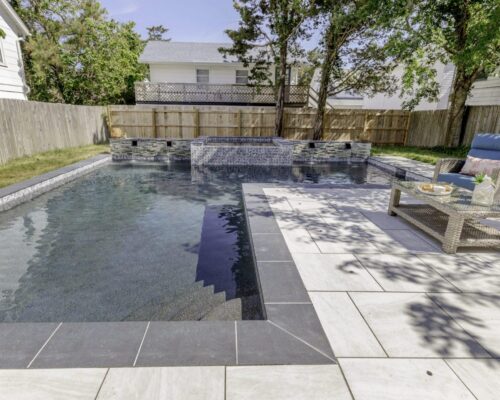 Rectangular backyard fiberglass pool with a raised custom spa and waterfall feature, surrounded by tile decking, lounge chair, and privacy fence with trees for enhanced outdoor living.
