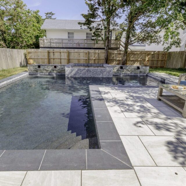 Rectangular backyard fiberglass pool with a raised custom spa and waterfall feature, surrounded by tile decking, lounge chair, and privacy fence with trees for enhanced outdoor living.
