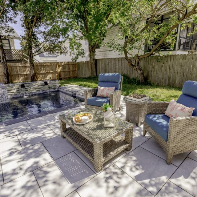 Wicker patio furniture with blue cushions and a glass coffee table set beside a luxury pool in a fenced backyard with trees and grass, perfect for stylish outdoor living.