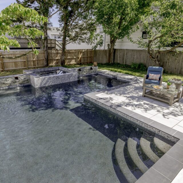 Rectangular luxury pool with attached custom spa, surrounded by a stone patio. Two cushioned lounge chairs and a small table sit on the deck, with trees and a wooden fence in the background.