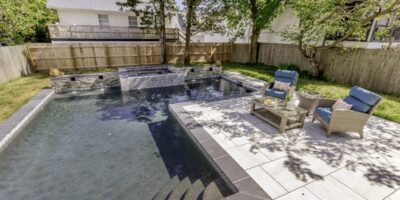 Backyard pool with a raised hot tub and custom spa, stone tile patio, wicker lounge chairs, small table, and shaded by trees, bordered by a wooden fence.
