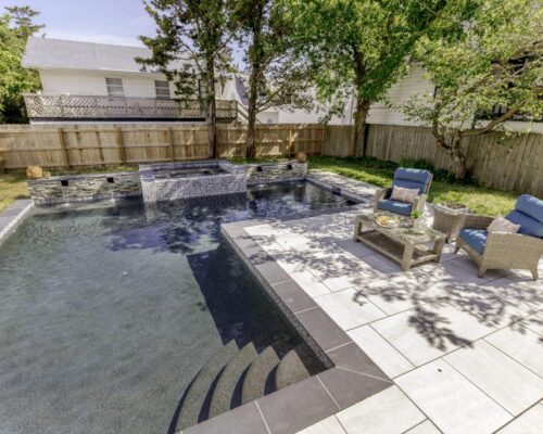 Backyard pool with a raised hot tub and custom spa, stone tile patio, wicker lounge chairs, small table, and shaded by trees, bordered by a wooden fence.