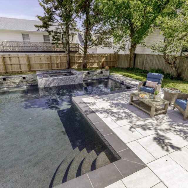Backyard pool with a raised hot tub and custom spa, stone tile patio, wicker lounge chairs, small table, and shaded by trees, bordered by a wooden fence.