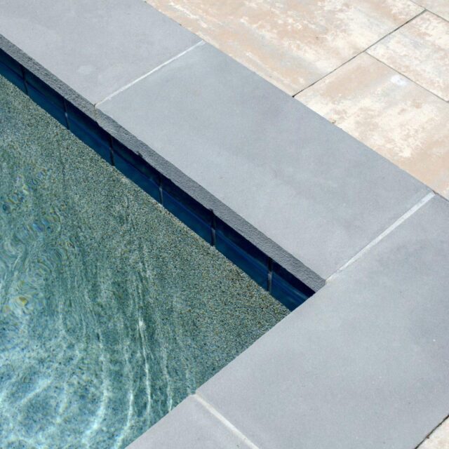 Close-up view of a luxury pool edge with clear water, gray coping stones, and beige tile paving surrounding the inground pool.