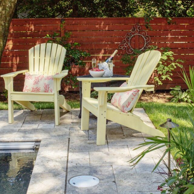Two yellow Adirondack chairs with patterned cushions sit by a fiberglass pool on a stone patio, next to a small table with a wine bucket, in a fenced backyard garden designed for relaxed outdoor living.