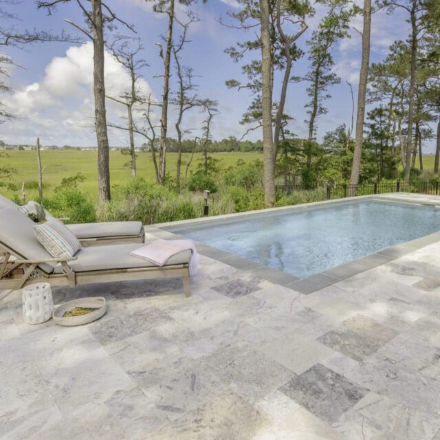 A rectangular outdoor swimming pool with a stone patio, two lounge chairs, and trees in the background overlooking open grassy fields—perfect for outdoor living.