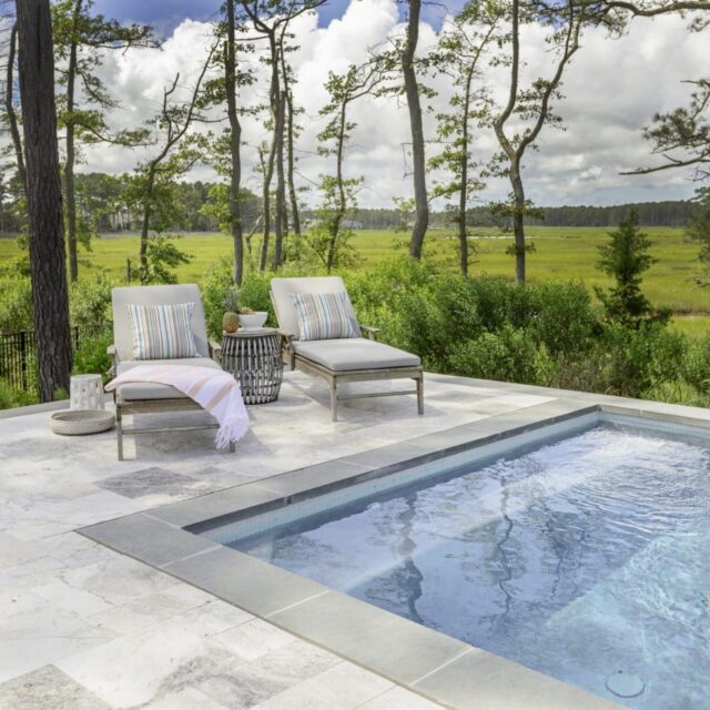 Two cushioned lounge chairs with striped pillows sit beside a clear inground pool, surrounded by greenery and trees under a partly cloudy sky.