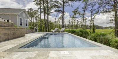 Rectangular luxury pool with stone tile decking, two lounge chairs, and a house nearby, nestled among trees and grassy landscape under a partly cloudy sky—a stunning outdoor living retreat.