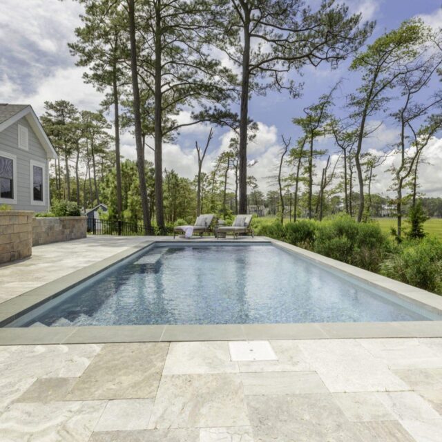Rectangular luxury pool with stone tile decking, two lounge chairs, and a house nearby, nestled among trees and grassy landscape under a partly cloudy sky—a stunning outdoor living retreat.