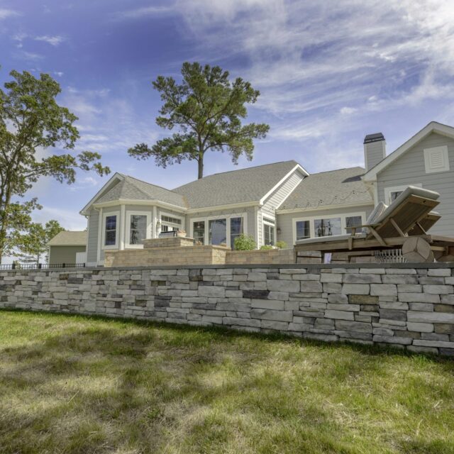 Single-story house with light siding, a stone retaining wall, and a patio area featuring lounge chairs and an inground pool in the backyard; surrounded by grass, tall trees, and a blue sky.