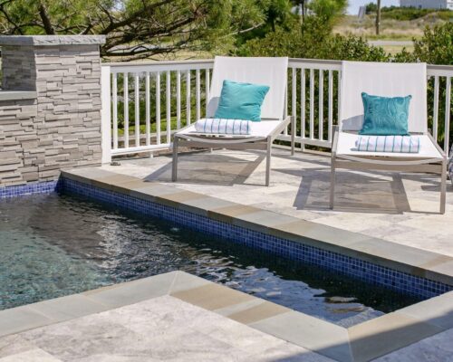 Two white lounge chairs with blue cushions and striped towels sit beside a small inground pool with a tiled deck and a white railing, creating a perfect luxury pools retreat.