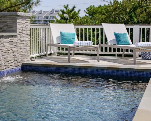 Two lounge chairs with pillows and rolled towels sit beside a luxury pool with a small stone waterfall feature and a white fence in the background, creating the perfect outdoor living retreat.
