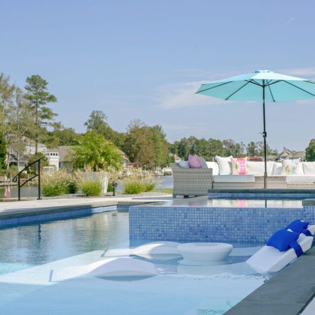 A modern backyard luxury pool with in-water loungers, a custom spa, blue umbrella, outdoor seating, and a view of a lake and houses in the background.