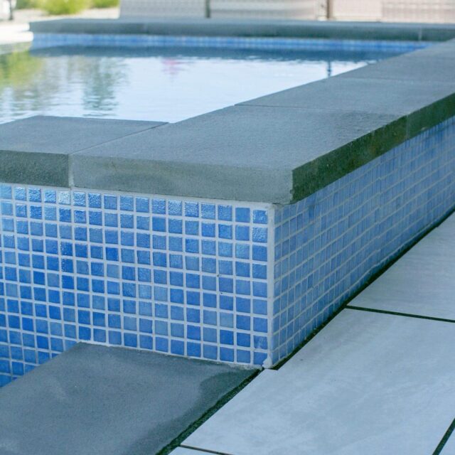 Close-up of a modern inground pool edge with blue square tiles and grey stone coping, beside white and grey tiled flooring—perfect for stylish outdoor living.