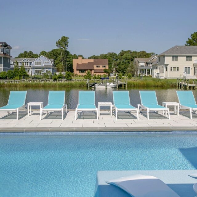 Poolside view with six blue lounge chairs on a patio, overlooking a canal with several houses and trees in the background under a clear sky—perfect for relaxing by fiberglass pools or custom spas.